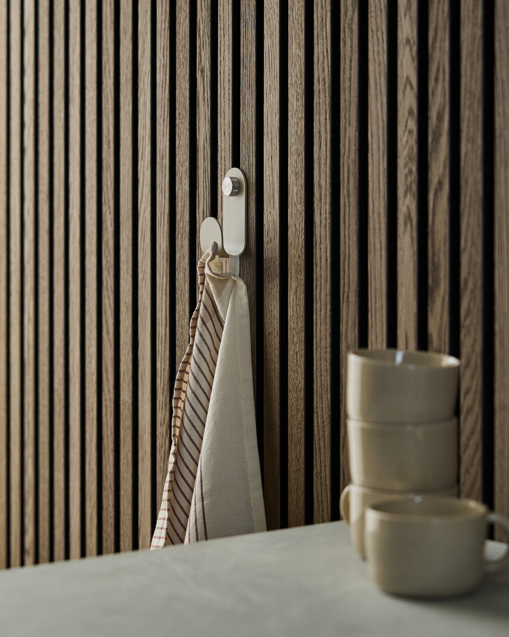 Et stribet håndklæde hænger på en WoodUpp Norr Single Hanger i beige, monteret på et akustisk lamelvægspanel i nordisk stil; tre beige krus er stablet foran.